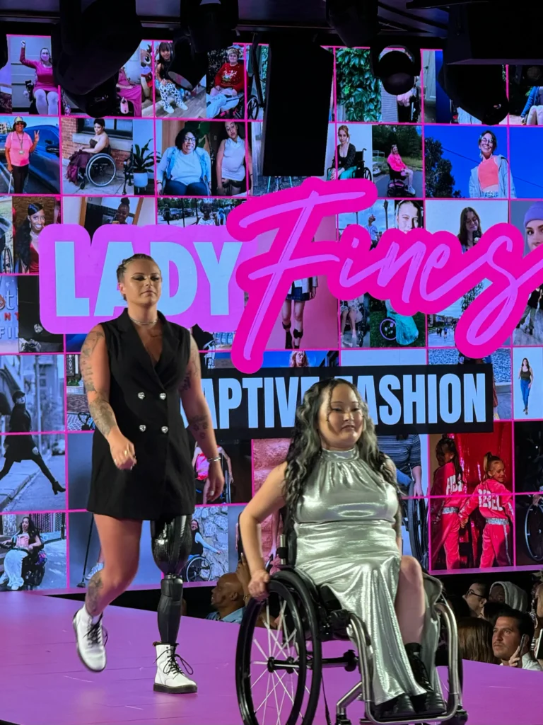 Two models walk the runway in a show for "LADY FINES ADAPTIVE FASHION," which is spelled out in large, bright pink neon lettering on the digital screen behind them. The model on the left, with dark hair and multiple tattoos, is walking and wearing a sleeveless, double-breasted black mini-dress, white lace-up boots, and a visible prosthetic leg below her left knee. The model on the right, with long dark hair, is sitting in a manual wheelchair and wearing a shimmering silver metallic halter-neck maxi dress. The background screen shows a collage of photos of diverse models with disabilities.