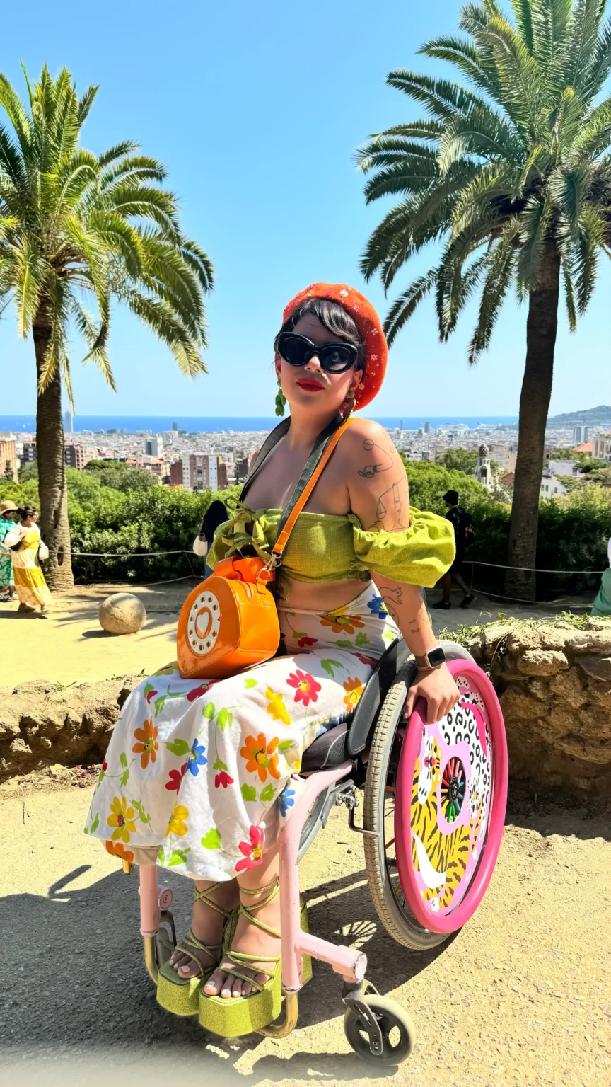 A brightly lit outdoor photo of a woman seated in a pink wheelchair at a scenic overlook, framed by two palm trees. She is wearing a green off-the-shoulder top, a white skirt with colorful flowers, green platform shoes, black sunglasses, and an orange beret. She has an orange rotary phone-shaped purse slung across her body. In the background, there is a view of a city sprawling below a clear blue sky.