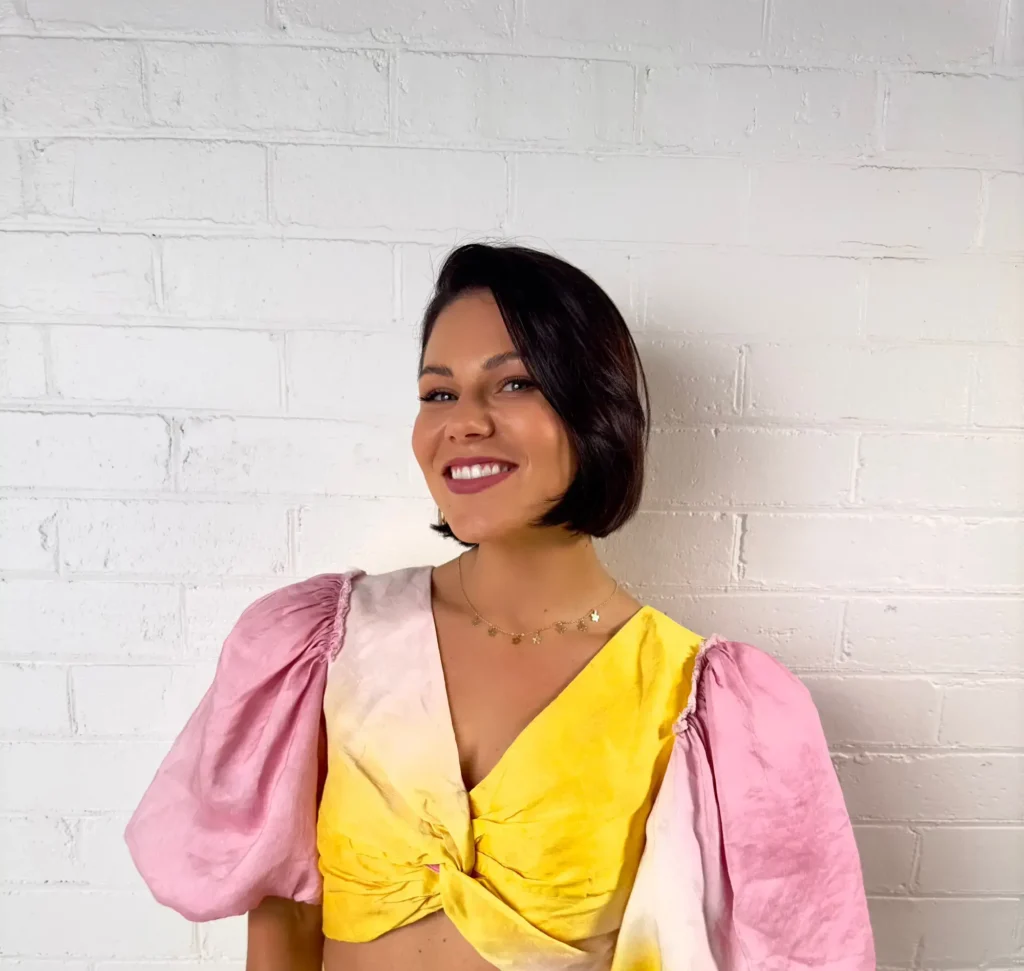 A headshot of a smiling woman with short, dark bobbed hair, wearing a brightly colored top. The top features large, voluminous sleeves and a knotted front, with a color gradient of pink, yellow, and white tie-dye. She is wearing a delicate gold star necklace and standing in front of a white painted brick wall.