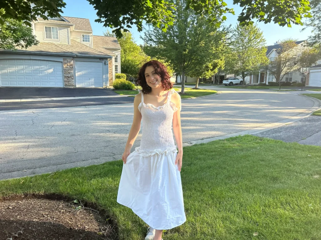 A young woman with short, reddish-brown curly hair smiles at the camera while standing in a suburban residential area. She is wearing a long, smocked, white summer dress with ruffles on the skirt. The setting is a paved street and grassy lawn with houses and garages in the background, under bright afternoon sunlight.