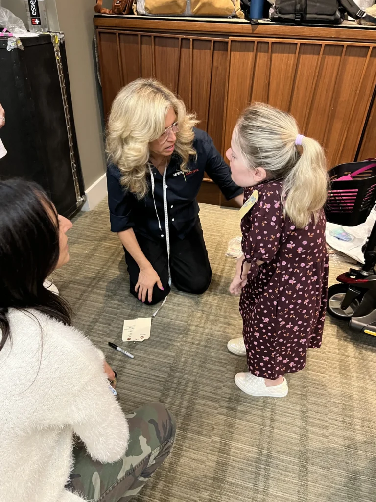 A group of people kneeling on a carpeted floor during a fitting. A blonde woman, likely Mindy Scheier, is wearing a dark shirt labeled 'SEWN ADAPTIVE' and is talking to a young girl with a high ponytail, who is standing in a maroon floral print dress. A tape measure is draped around the woman's shoulder, and another person is seated nearby.