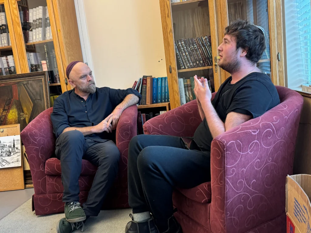 Jacob and Rabbi Eli sit in plush, maroon armchairs during a conversation at Congregation Emanu-El. Rabbi Eli, on the left, is a middle-aged man with a grey beard and a purple kippah, listening intently. Jacob, on the right, is seen in profile wearing a black t-shirt and a kippah, gesturing with his hands as he speaks. The background features tall wooden bookshelves filled with various religious texts.