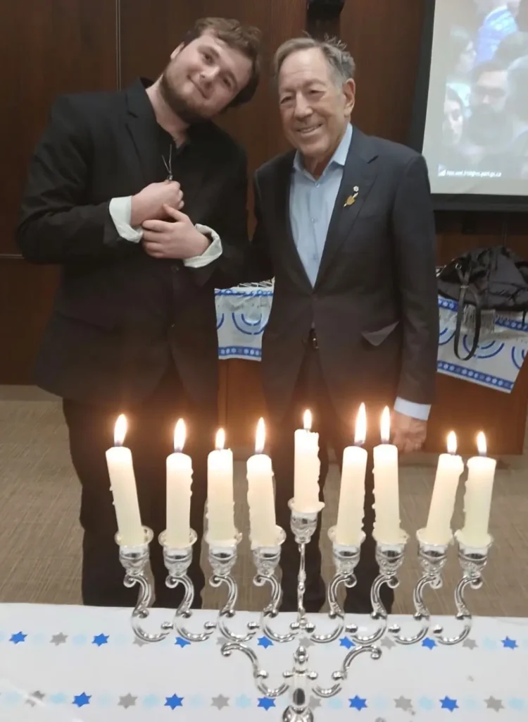 A medium shot of two men standing together behind a silver Chanukiah (menorah) with nine white candles lit. On the right, the Honorable Irwin Cotler, an older man with grey hair, wears a dark suit and a light blue shirt. On the left, Jacob, a younger man with a beard and dark hair, wears a dark blazer and leans his head toward Mr. Cotler. The setting is an indoor room with wood-paneled walls and a table decorated with a blue and white Star of David runner.