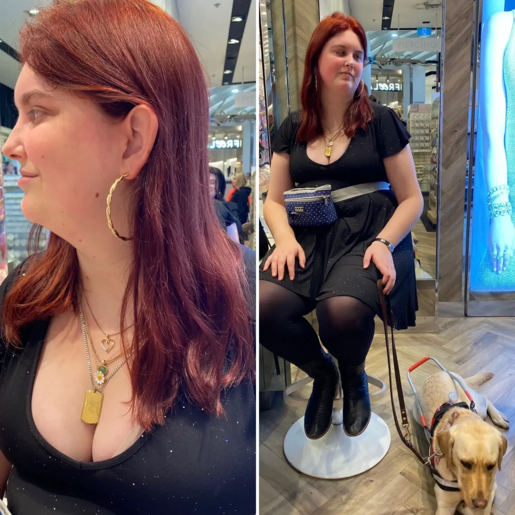 A two-panel photo of a young woman with long reddish-brown hair. The left panel is a close-up profile view of her face and chest, focusing on a large gold hoop earring and several layered necklaces, including a gold pendant and a necklace with a heart and a green stone. The right panel is a full shot of her sitting on a white stool in a retail store, wearing a black dress, black tights, black boots, and a small black and white patterned waist bag. A guide dog is lying patiently at her feet.