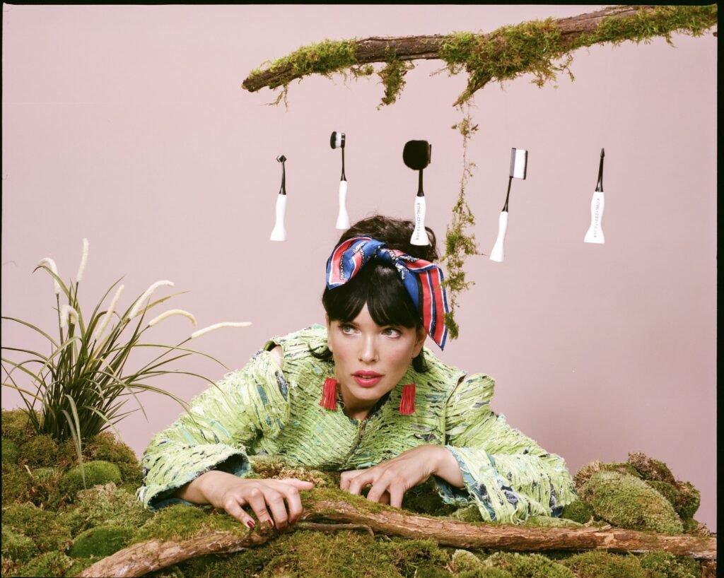 A woman with dark hair and bangs, wearing a vibrant green textured jacket and a striped headscarf, peers out from a landscape of moss and natural elements. Above her, five makeup brushes with white handles and black heads are suspended, referencing the Khol Kreatives brand.