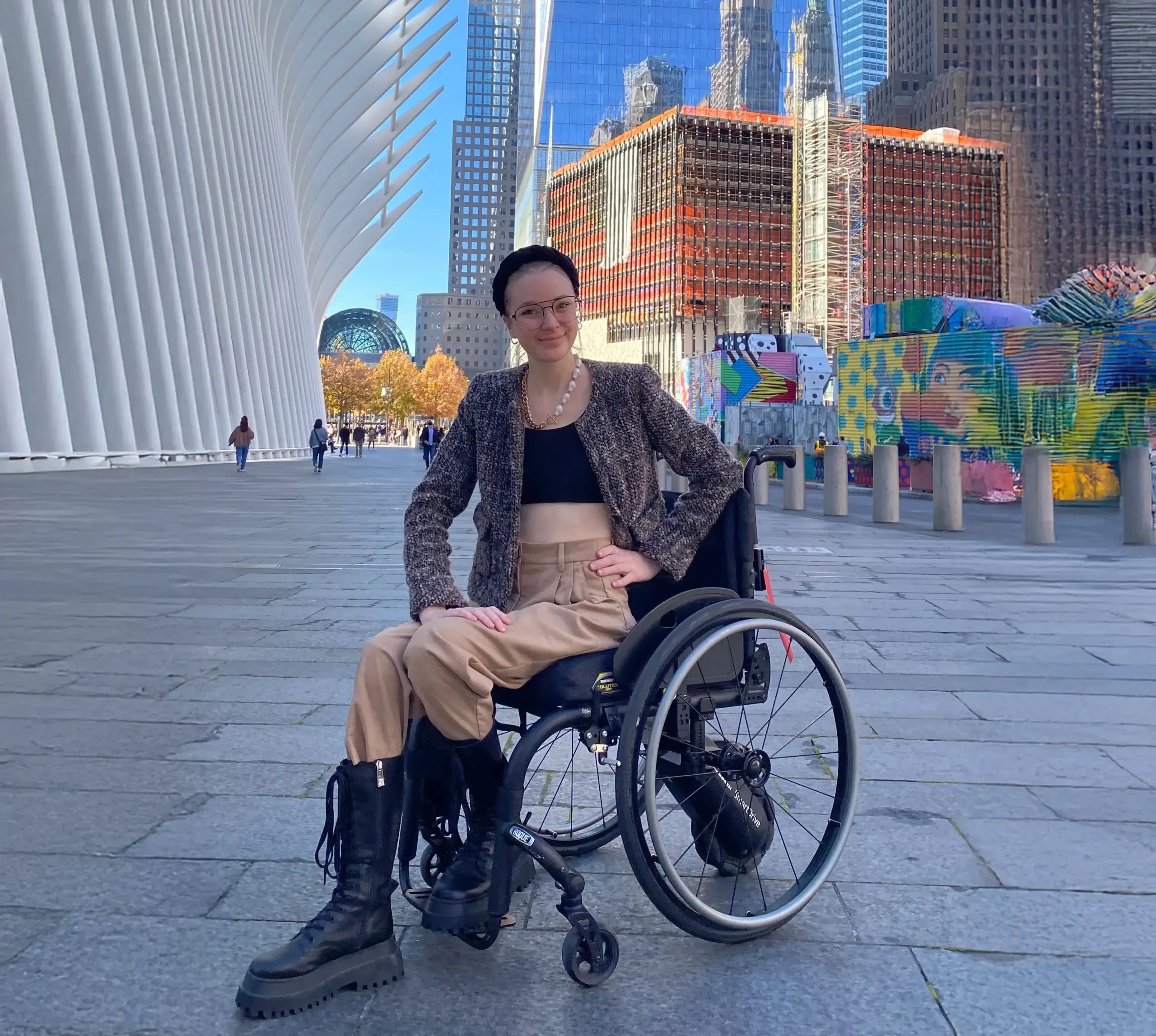 A young woman in a manual wheelchair posing outdoors in a plaza with modern architecture. She is dressed in stylish fall attire: a black crop top, light beige trousers, a dark brown tweed jacket, a chunky pearl necklace, a black headband, and black combat boots. In the background are the curved white structure of the Oculus and surrounding skyscrapers in New York City.