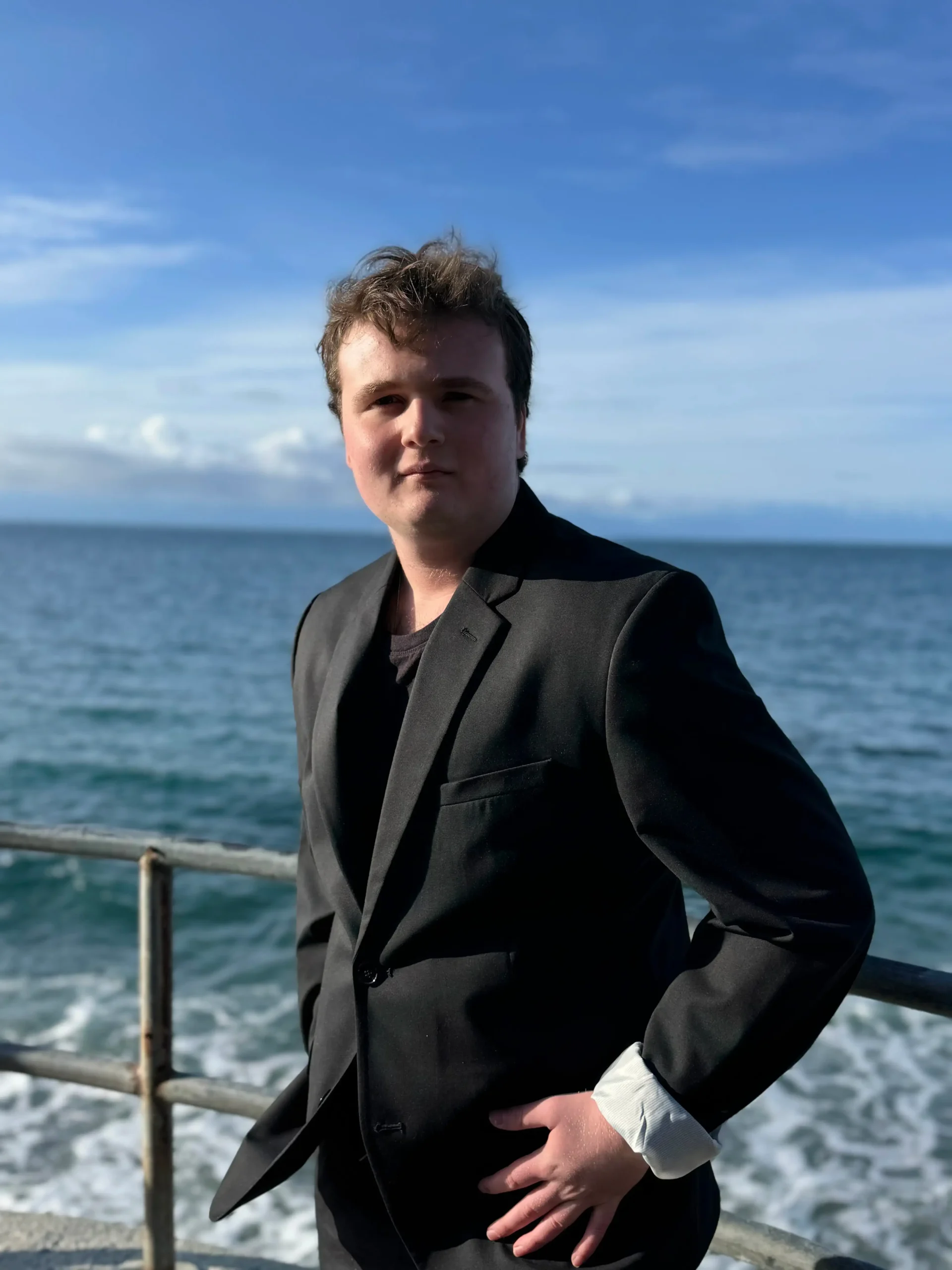 A young man poses on a pier or seawall, looking directly at the camera. He is wearing a black suit jacket over a black shirt, with his left hand on his hip, and the jacket cuffs rolled up to expose white lining. The blue ocean and a partially cloudy sky are visible behind him.
