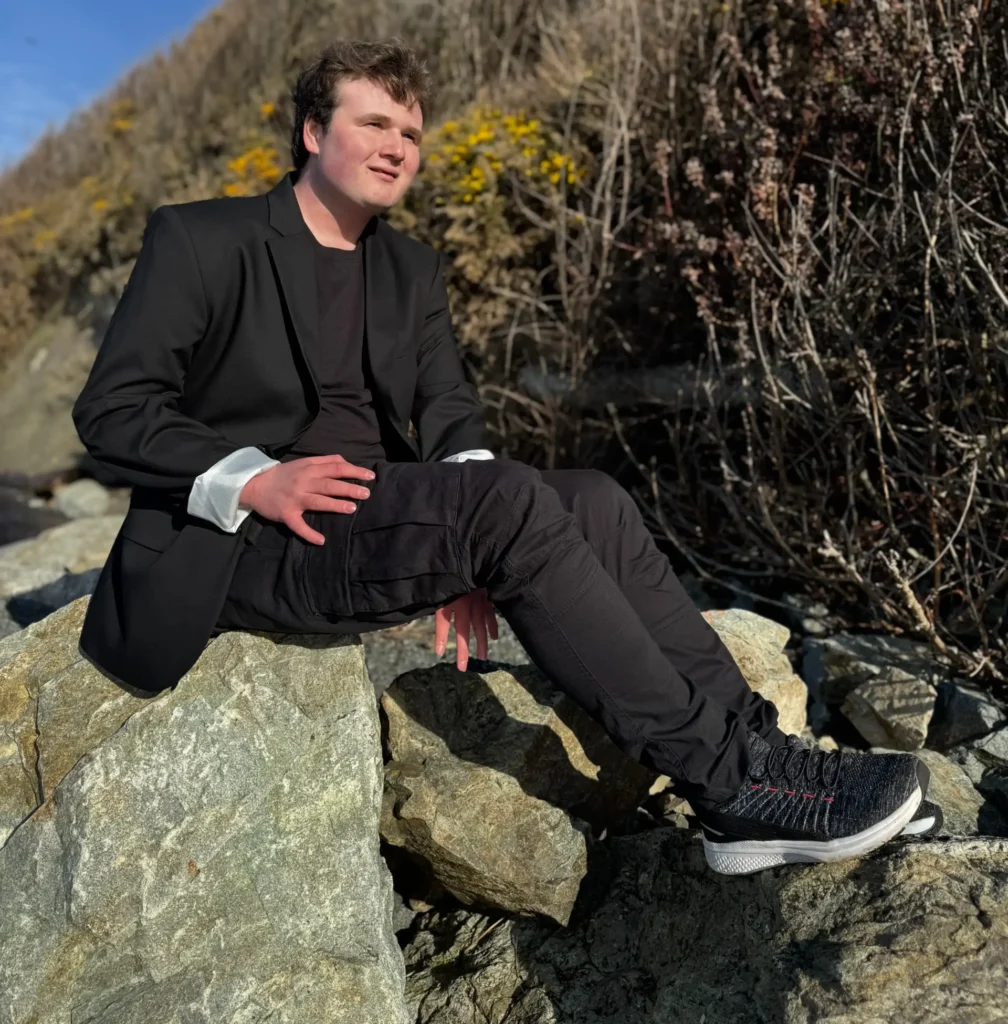 A young man sits on large gray rocks near the ocean, looking off to the side. He is wearing a black suit jacket, black t-shirt, and black cargo pants with sneakers. His jacket cuffs are rolled up. Behind him is a steep, dry embankment covered in scrub brush with patches of yellow flowers, under a bright blue sky.