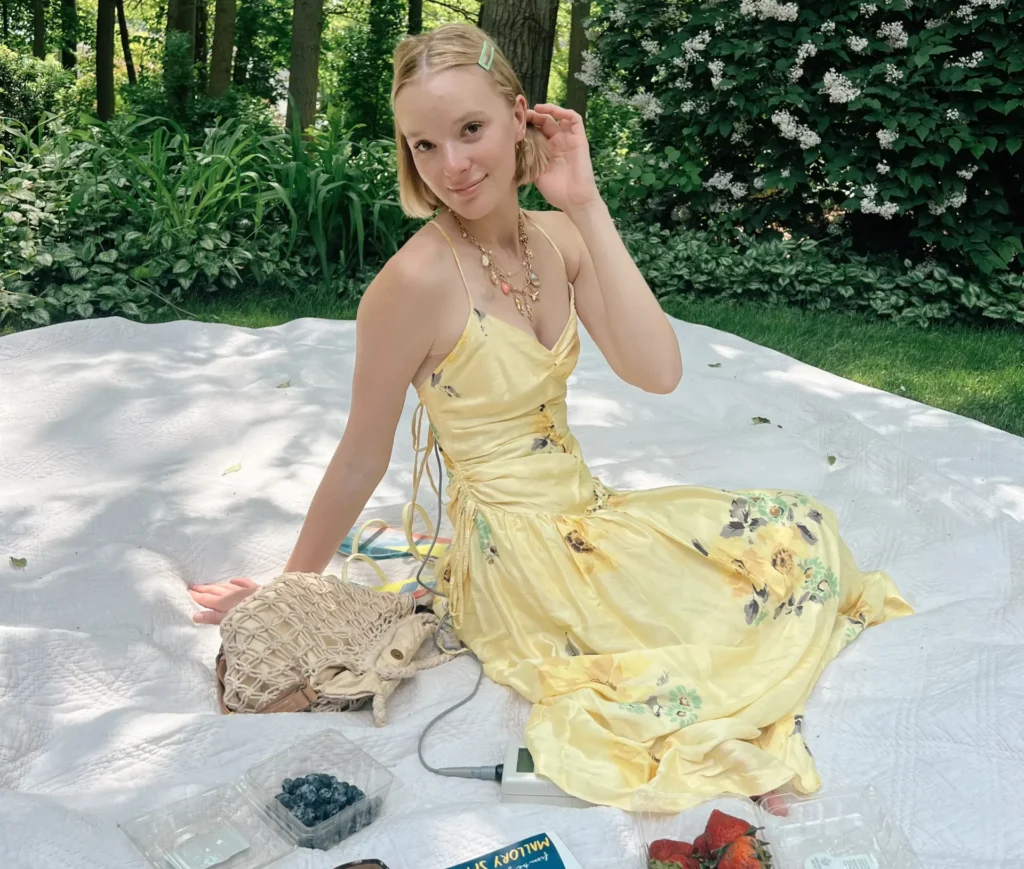 A young woman sitting on a white picnic blanket outdoors in a lush, green yard surrounded by trees and bushes. She is wearing a bright yellow sundress with a floral print. In front of her are a book titled "DIARY OF A DYING GIRL," a woven bag, sunglasses, and plastic containers with blueberries and strawberries.
