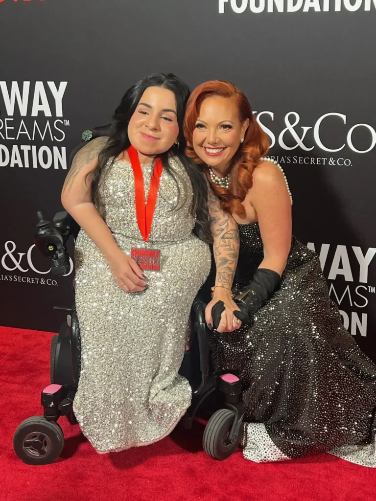 A red carpet photo of two smiling women posing together. On the left, a woman in a power wheelchair wears a shimmering silver sequined gown and a red medal around her neck. On the right, a woman with red hair in a black sequined gown kneels slightly to hold her hand. The background displays the logos for 'Runway of Dreams Foundation' and 'VS&Co'.