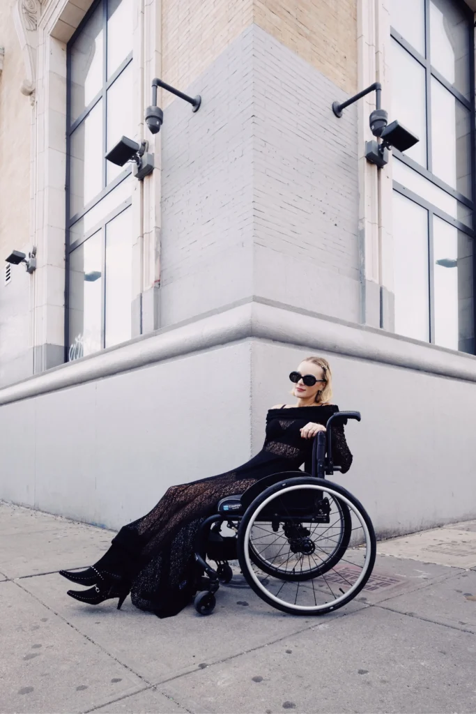 A stylish young woman sitting in a manual wheelchair on a city sidewalk. She is wearing a long, sheer black lace gown with off-the-shoulder details, black pointed boots with studded details, and large black sunglasses. She is positioned against the corner of a light-colored, multi-story building with security cameras.