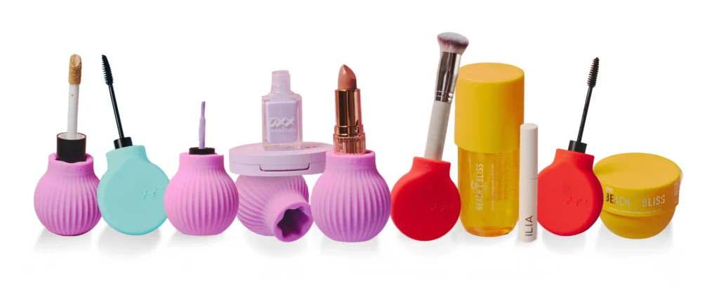 A row of various makeup and skincare products set against a white background. Many of the products feature distinctive, ribbed, bulb-shaped silicone handles in neon pink, turquoise, and red, holding items like a gold liquid eyeshadow wand, mascara, and a foundation sponge. Other products include a lavender nail polish, a nude lipstick in a gold tube, a large yellow oil dropper bottle labeled "BEACH BLISS," an ILIA tube, and a yellow tub of "BEACH BLISS" body butter.