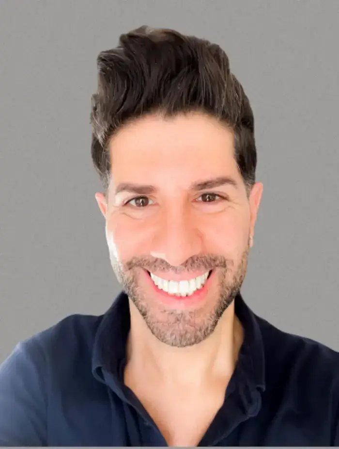 A close-up portrait of Ramy Gafni, a dark-haired man with a friendly smile, clean white teeth, and dark eyes, wearing a navy blue collared shirt. He has styled dark hair and a neatly trimmed goatee/mustache. The background is a plain, uniform gray.