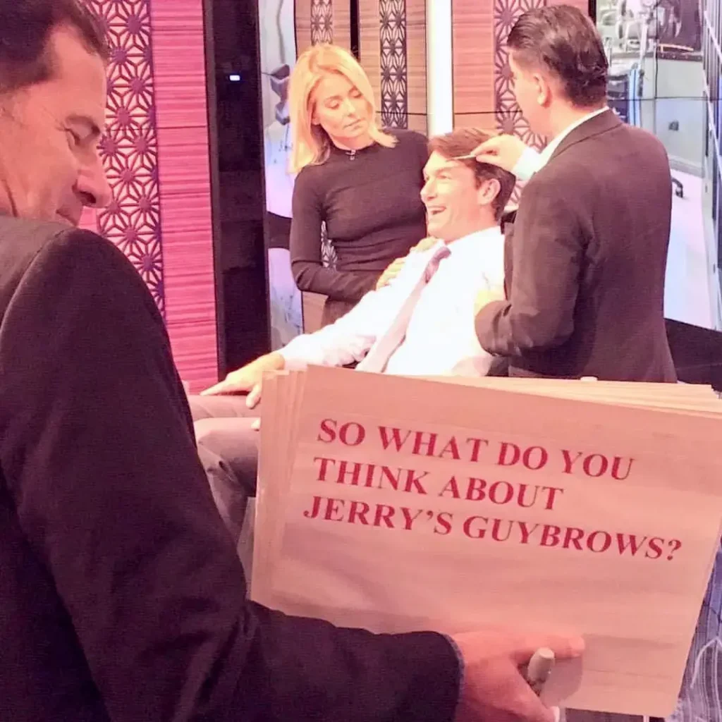 A man, possibly Jerry Seinfeld, is seated in a makeup chair while another man (likely a makeup artist) works on his eyebrows. A woman (likely Kelly Ripa) stands behind him, looking on. In the foreground, a man in a dark suit holds up a large sign that reads in red text: "SO WHAT DO YOU THINK ABOUT JERRY'S GUYBROWS?". The setting appears to be a brightly lit television studio set.