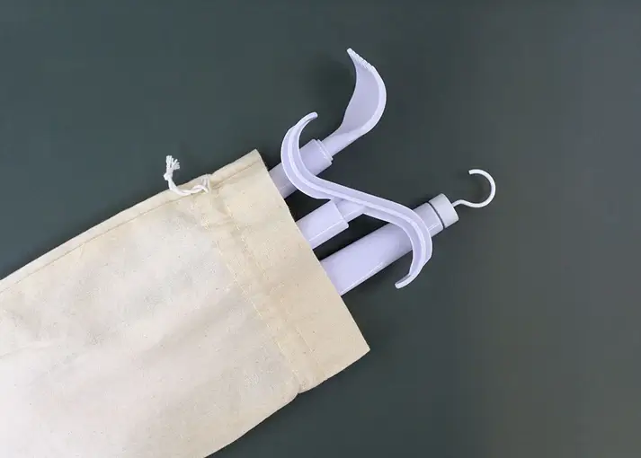 A light beige canvas drawstring bag is shown against a dark greenish-gray background. Peeking out of the bag are the white plastic handles of two dressing aids: a long-handled shoehorn or hook and a garment buttoning hook or zipper pull.