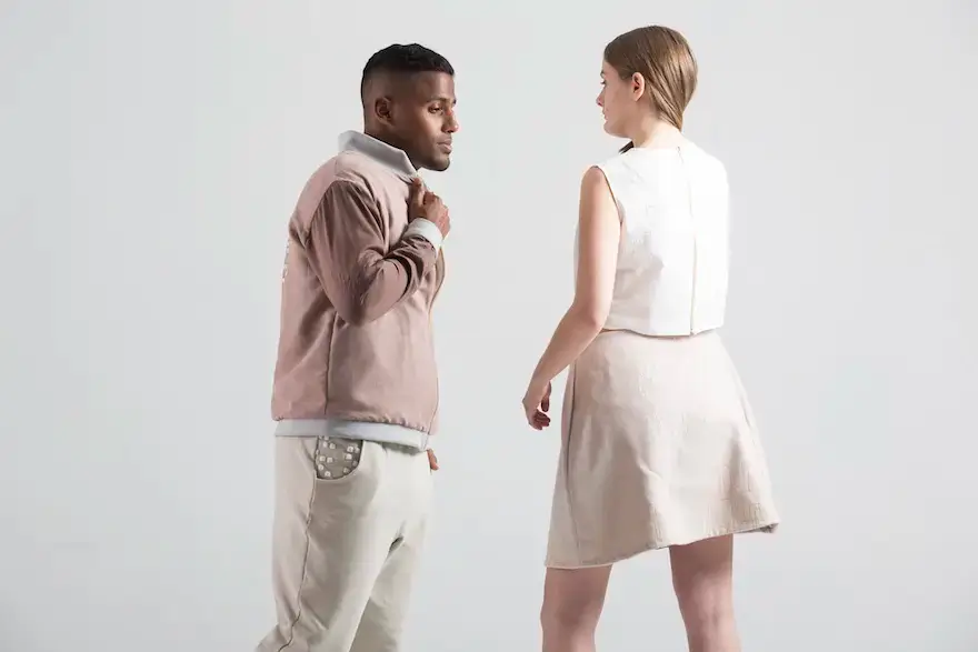 Two models standing facing each other against a plain white background. The man on the left wears a light brownish-pink bomber jacket over a white top and light khaki pants, with a pocket detail on the pants featuring metallic studs or raised dots. The woman on the right wears a white sleeveless crop top and a plain, light tan, A-line skirt.
