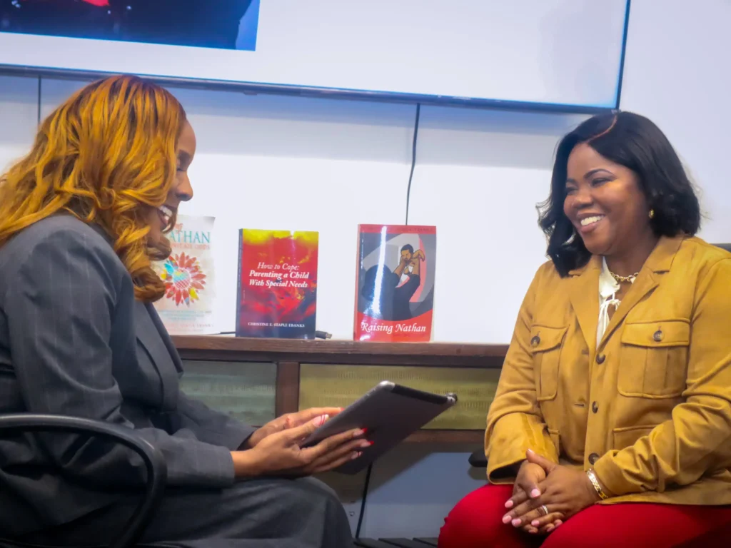 Two women are seated and smiling during a discussion in an office setting. Three books about parenting children with special needs are displayed on a shelf behind them. One woman holds a tablet while the other listens attentively.