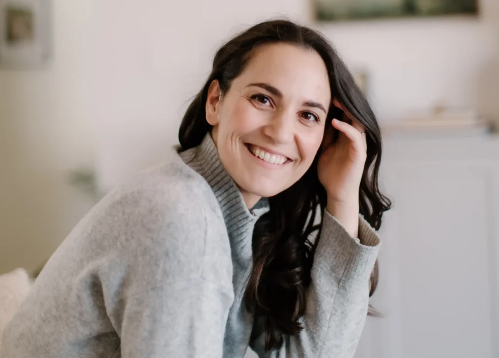 A portrait of a smiling woman with long, dark wavy hair wearing a grey turtleneck sweater. She is leaning forward slightly with one hand tucked behind her ear, sitting in a bright, minimalist room with a blurred artwork in the background.