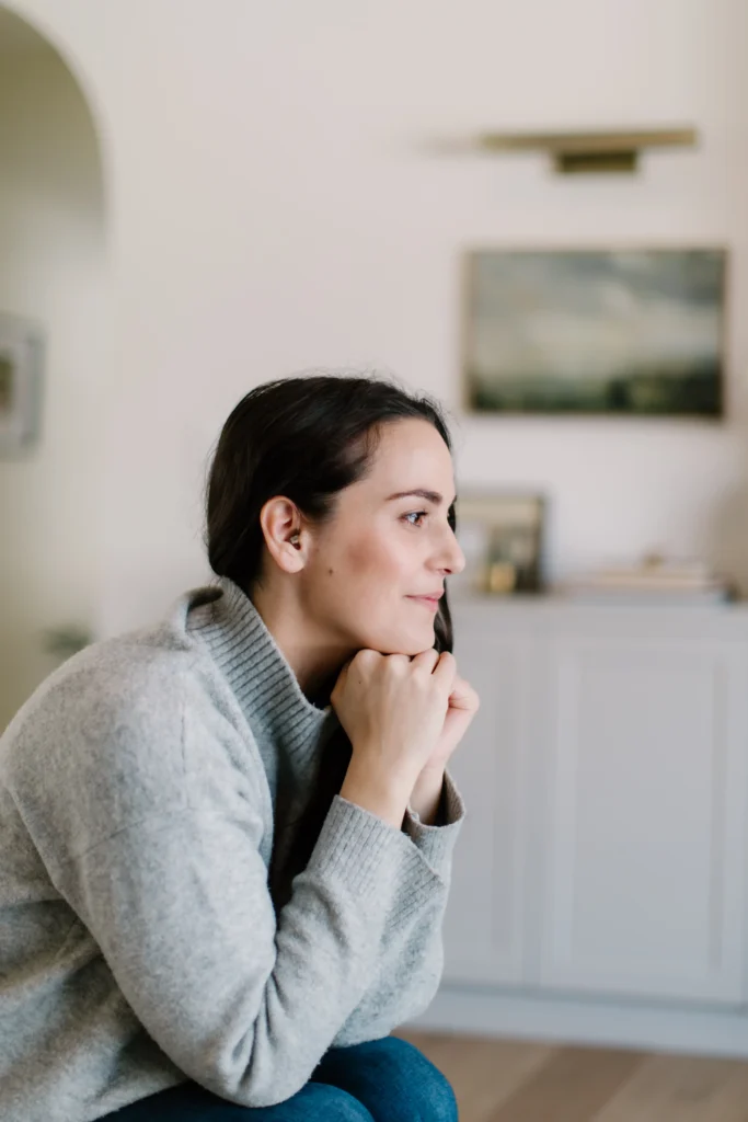 A profile shot of a woman with dark hair tucked behind her ear, revealing an in-ear hearing aid or earplug. She is resting her chin on her folded hands, looking thoughtfully off-camera in a well-lit indoor setting.