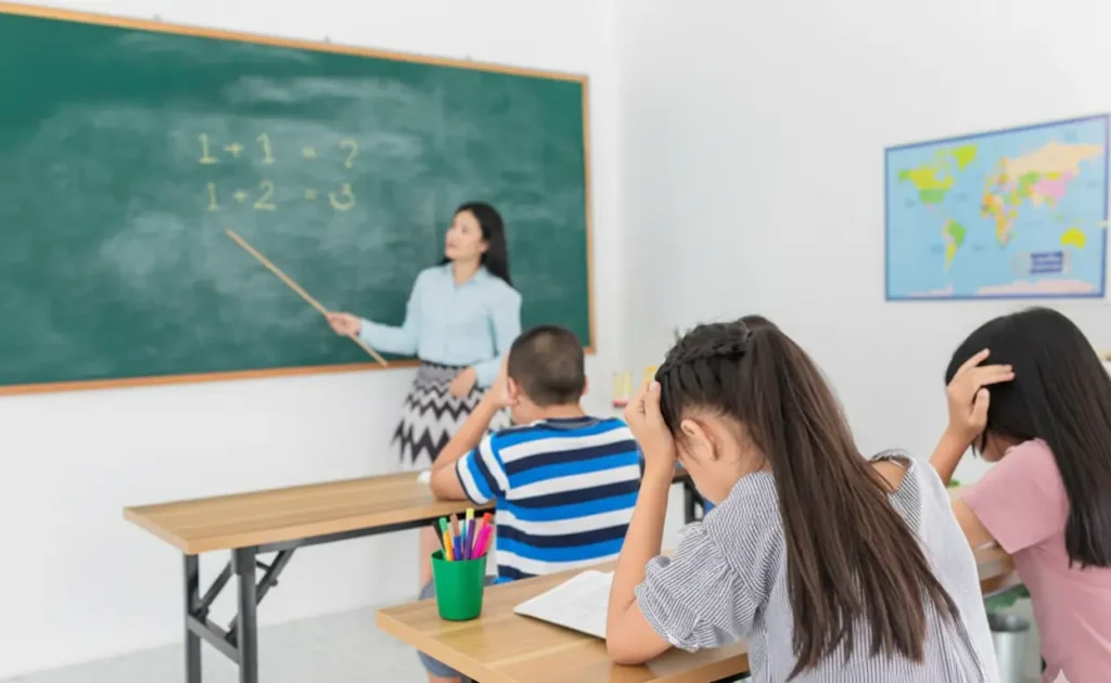 A classroom scene where a teacher stands at a green chalkboard pointing to simple addition equations. In the foreground, three students are seen from behind, sitting at their desks with their heads in their hands, appearing frustrated or tired.