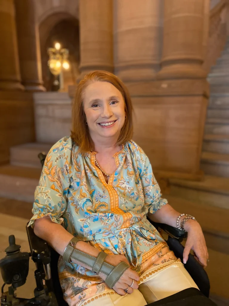 A full-body portrait of a smiling woman with reddish hair sitting in a power wheelchair inside a building with ornate stone architecture and a grand staircase. She is wearing a light teal floral print blouse with orange accents and white trousers. Her right hand is supported by a green medical splint.