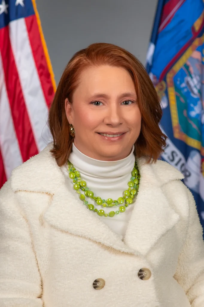 A formal professional portrait of a woman with shoulder-length reddish-brown hair. She is wearing a white textured coat over a white turtleneck, accented by a multi-strand lime green bead necklace. She stands in front of the United States flag and the New York State flag.