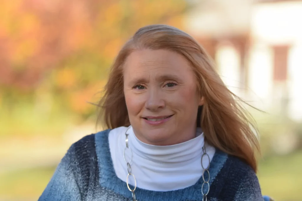 A close-up outdoor headshot of a woman with long, strawberry-blonde hair blowing slightly in the wind. She is smiling gently and wearing a blue marled sweater over a white turtleneck, accessorized with a silver link necklace. The background is a soft, out-of-focus autumn landscape with orange and green tones.