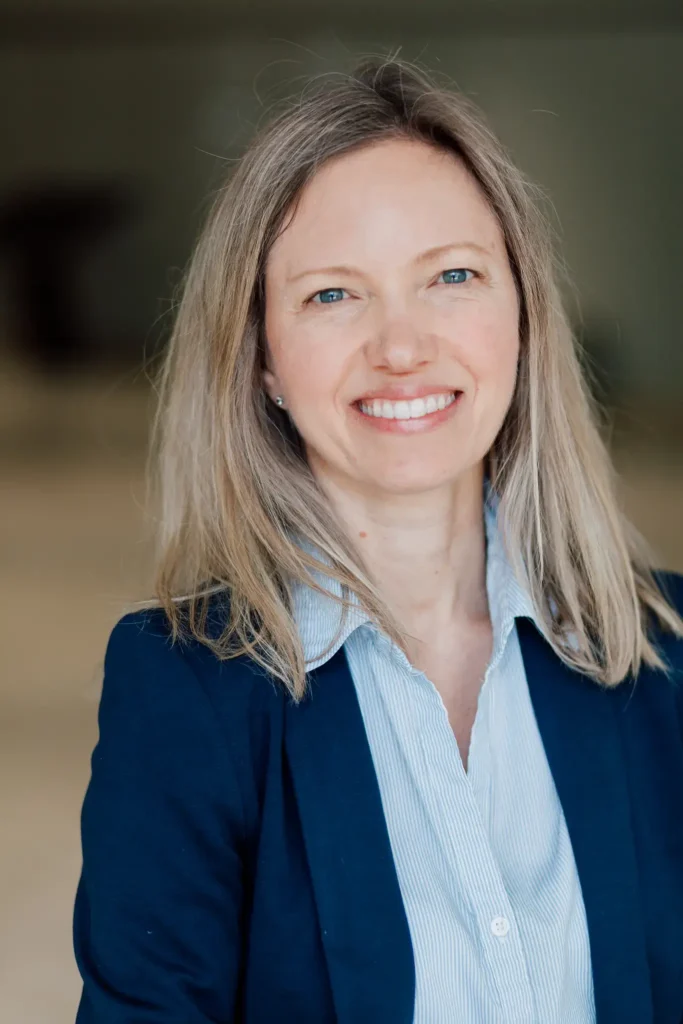 A professional headshot of a woman with long blonde hair and blue eyes. She is smiling warmly at the camera, wearing a navy blue blazer over a light blue and white striped button-down shirt. The background is soft-focus and neutral.