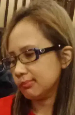 A close-up headshot of an Asian woman with long, brownish-blonde hair wearing rectangular black-framed glasses. She is looking slightly downward with a neutral expression, wearing a red top.