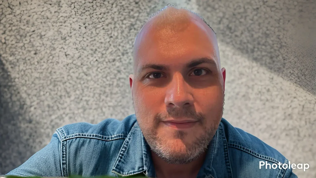 A close-up headshot of a man with a shaved head and light stubble, smiling warmly at the camera. He is wearing a blue denim shirt. The background is a textured, light-colored wall with a subtle dappled light effect.