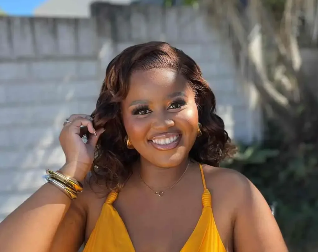 A bright, cheerful photo of the same woman sitting in her wheelchair, smiling radiantly at the camera. She is wearing a vibrant, silky yellow halter-style dress and gold jewelry, with her wavy hair framing her face. The background shows a sunny outdoor area with a white brick wall and palm trees.