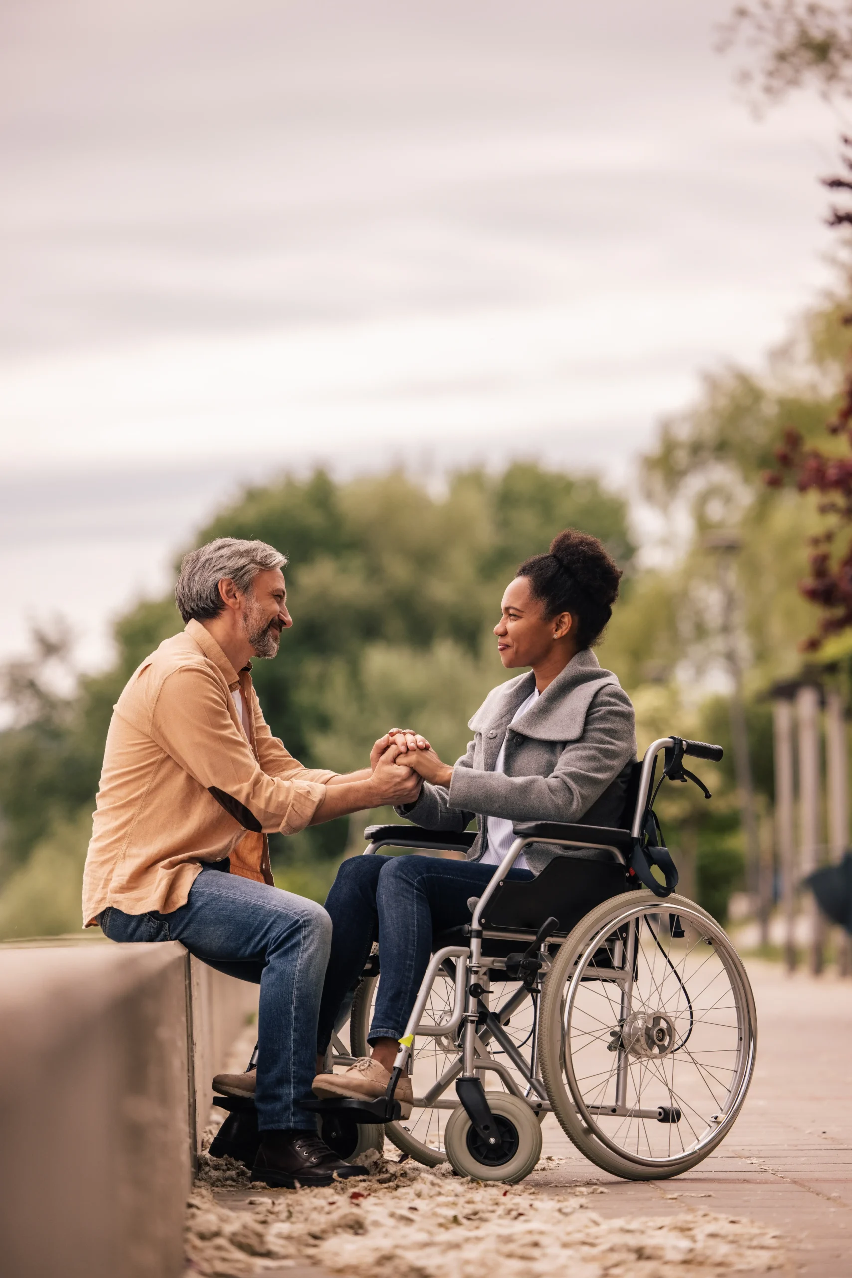 A middle-aged man with salt-and-pepper hair sits on a low stone wall outdoors, tenderly holding both hands of a Black woman sitting in a wheelchair. They are looking into each other's eyes and smiling warmly against a soft-focus background of green trees and a pale sky.