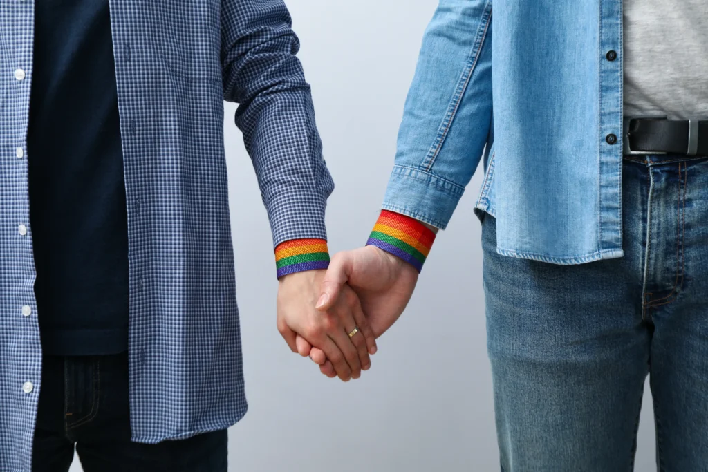 A close-up shot of two people standing side-by-side, holding hands. Both individuals are wearing rainbow-striped wristbands over the cuffs of their long-sleeved shirts (one blue checkered, one denim). The background is a neutral, light gray.