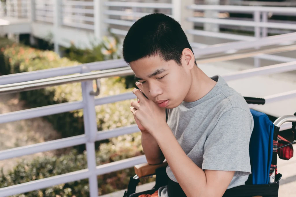 A young Asian boy with a short buzz cut sits in a blue wheelchair on an outdoor walkway. He is wearing a grey t-shirt and has his eyes closed, resting his head slightly against his hand in a pensive or resting pose. Metal railings and green foliage are visible in the background.