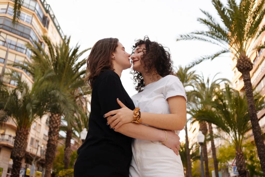 Two young women in a loving embrace on a sunny city street lined with tall palm trees and multi-story buildings. One woman wears a black shirt and the other a white t-shirt; they are leaning in close with their eyes closed, smiling gently as their faces almost touch.