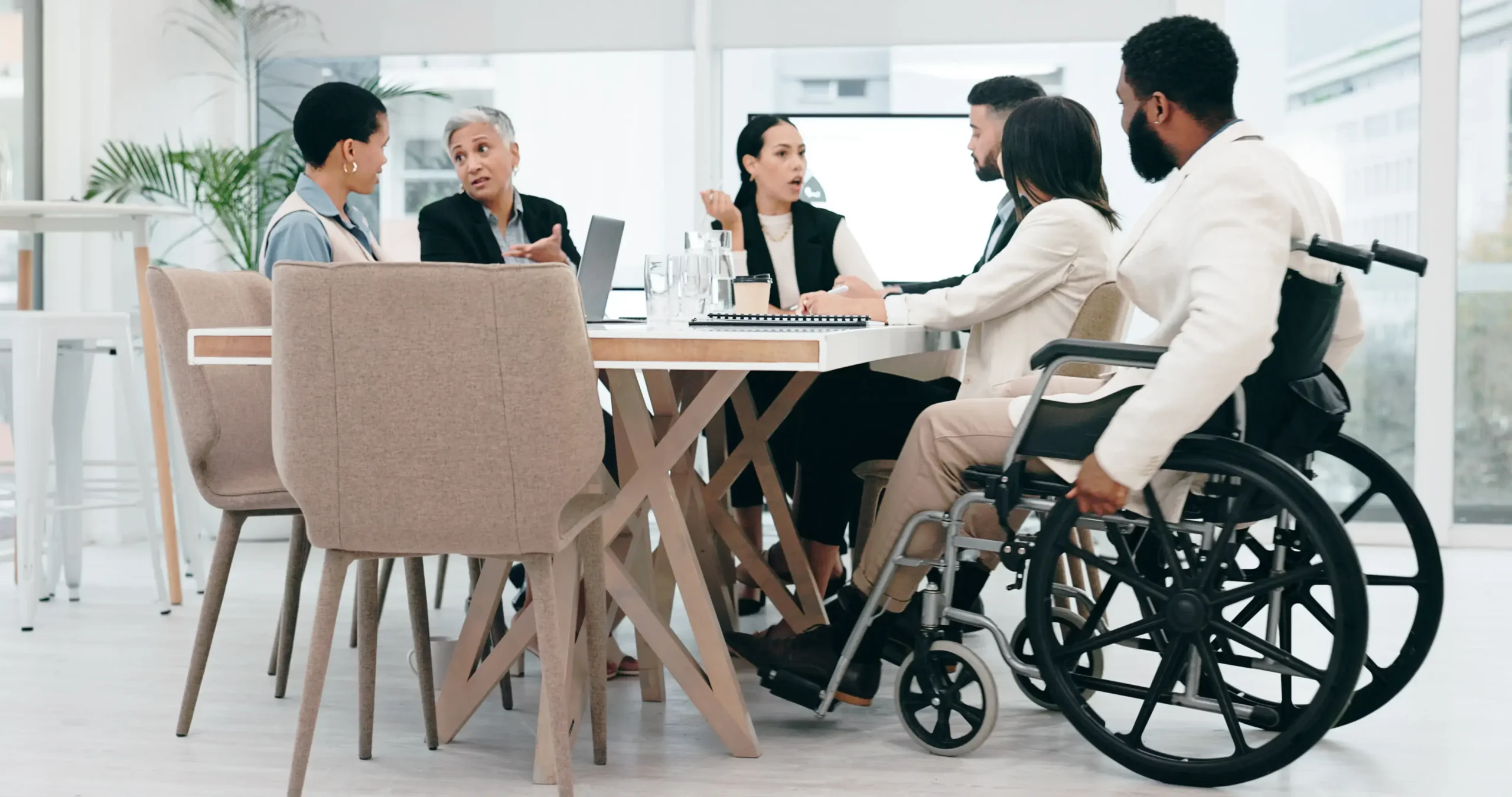 A diverse group of colleagues engaged in an active discussion around a large white conference table in a modern, bright office. The group includes people of various ethnicities and ages, including a man in the foreground using a black wheelchair.