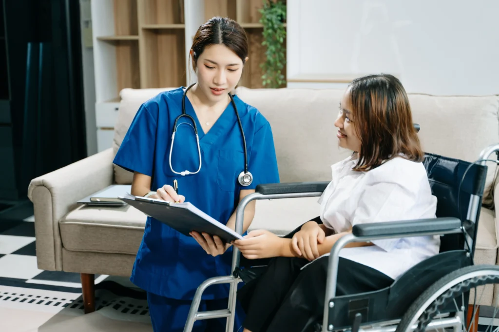 A nurse in blue medical scrubs with a stethoscope around her neck stands next to a young woman in a wheelchair. The nurse is writing on a clipboard while the woman looks up at her with a slight smile. They are in a modern, brightly lit indoor living space.