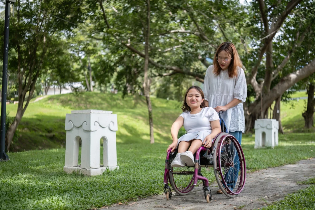 A young woman with a disability sits in a purple manual wheelchair on a paved path in a lush green park. Another woman in a white blouse stands behind her, holding the wheelchair handles as they enjoy a walk together outdoors.