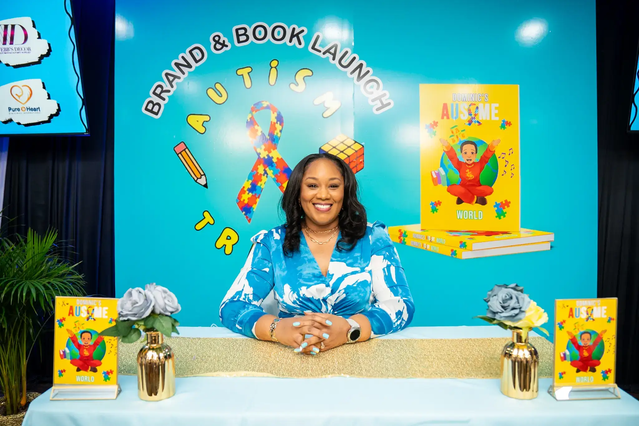 Author Giselle Trail McIntosh smiling behind a display table at the Autism Trails Brand and Book Launch, featuring copies of Dominic's Ausome World and floral arrangements.