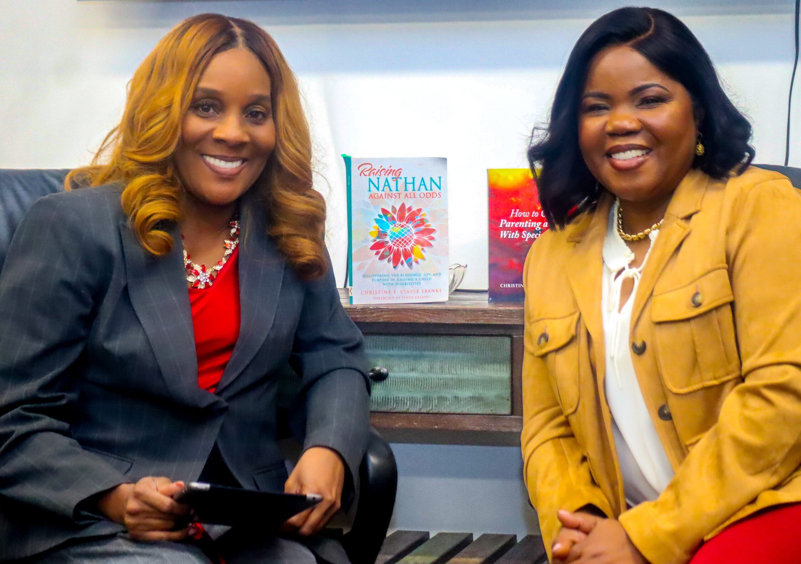 Two women are sitting together indoors, smiling for the camera. The woman on the left has long blonde wavy hair and wears a dark blazer over a red top. The woman on the right has dark hair and wears a tan jacket over a white blouse. Between them on a table is a book titled "Raising Nathan Against All Odds" featuring a colorful sunflower on the cover.