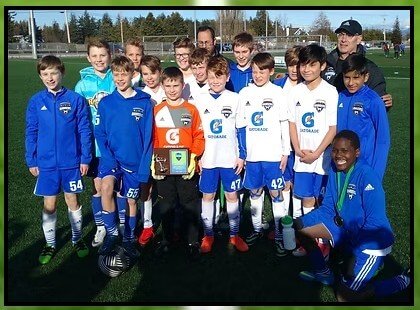 A youth soccer team in blue and white uniforms poses for a group photo on a sunny turf field. One player in the center holds a championship trophy, and the players are smiling, some wearing medals. Two adult coaches stand in the back row, and a line of evergreen trees is visible behind the field fence.