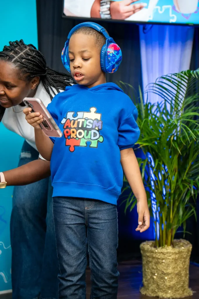 A young boy named Dominic wearing blue noise-canceling headphones and an "Autism Proud" sweatshirt, looking at a phone while standing in his personal space at the Autism Trails event.