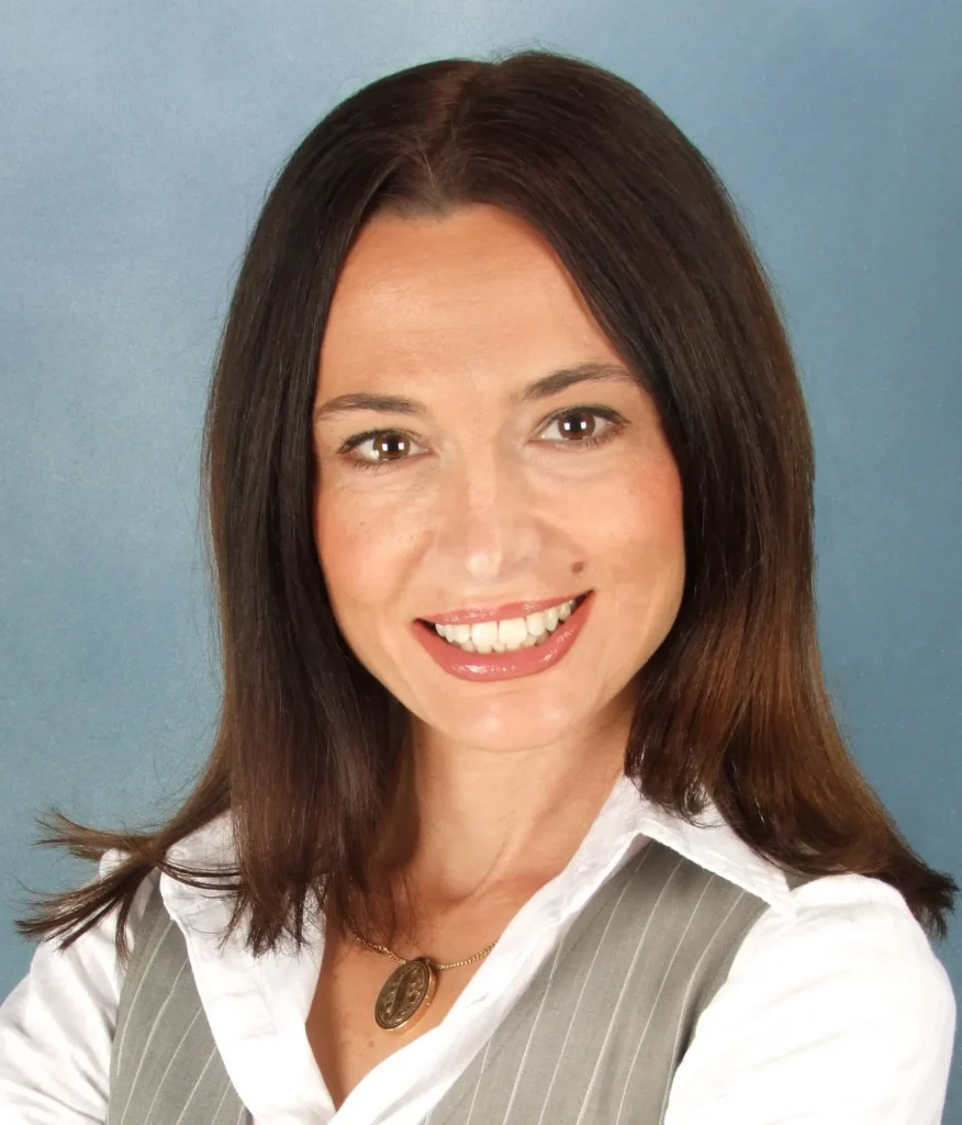 A professional headshot of a woman with long, dark brown hair and brown eyes, smiling at the camera. She is wearing a white collared shirt under a gray pinstriped vest and a gold circular pendant necklace. The background is a solid, textured blue.