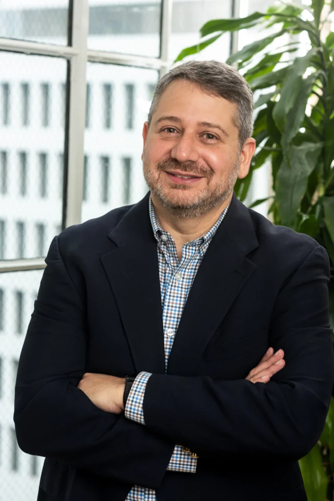 Joe Devon, co-founder of Global Accessibility Awareness Day, smiling with arms crossed in a professional office setting with a window and green plant.