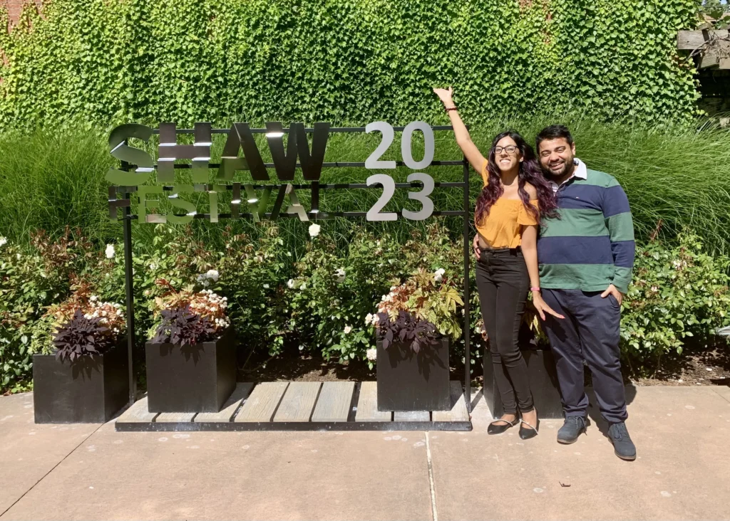 A bright, outdoor photo of a couple standing next to a "SHAW FESTIVAL 2023" sign. The woman, in a yellow crop top and black jeans, has one arm raised in a celebratory gesture. The man, in a green and navy striped rugby shirt, stands beside her with a hand in his pocket. They are positioned in front of a dense wall of green ivy and several black planters filled with flowers.