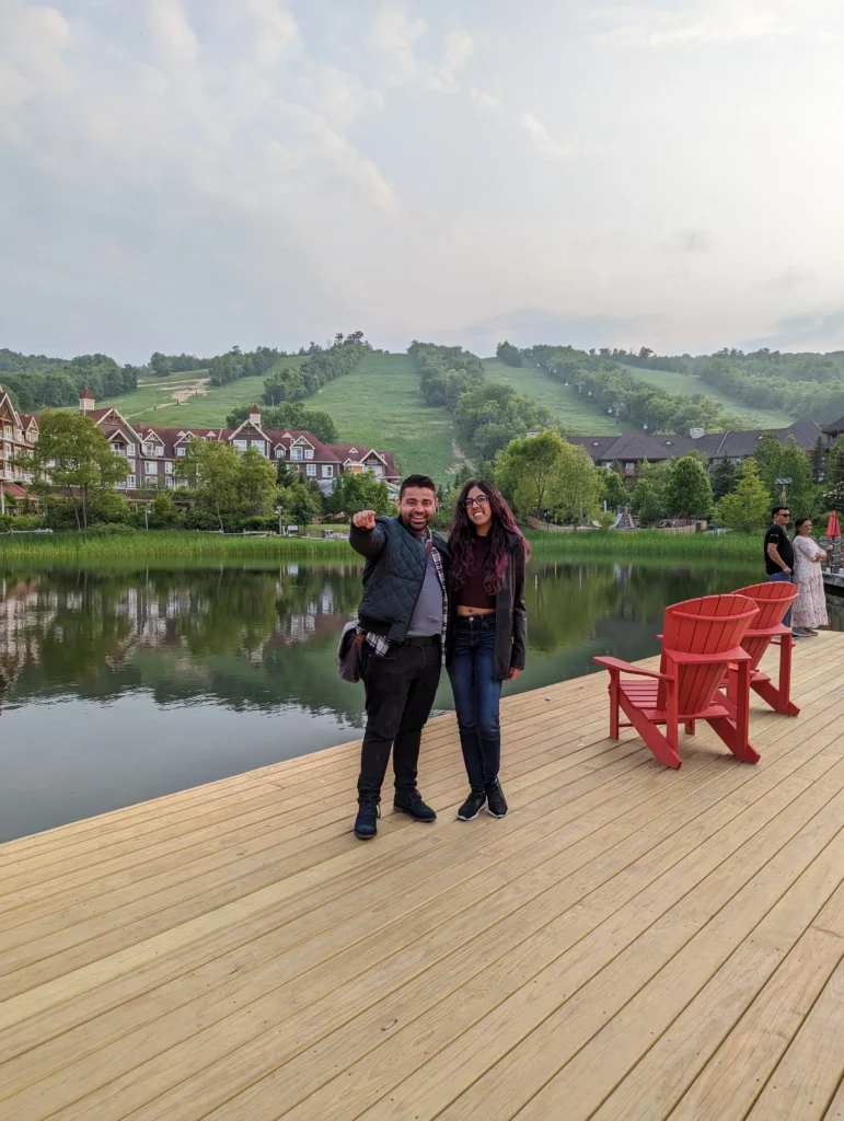 A smiling couple stands on a light-colored wooden dock overlooking a calm lake. The man, on the left, wears a quilted navy jacket and black pants, pointing toward the camera. The woman, on the right, has long dark hair with purple tints and wears a dark jacket, maroon top, and jeans. In the background, lush green hills with ski runs are visible under a soft, overcast sky, with resort-style buildings and two red Adirondack chairs nearby.