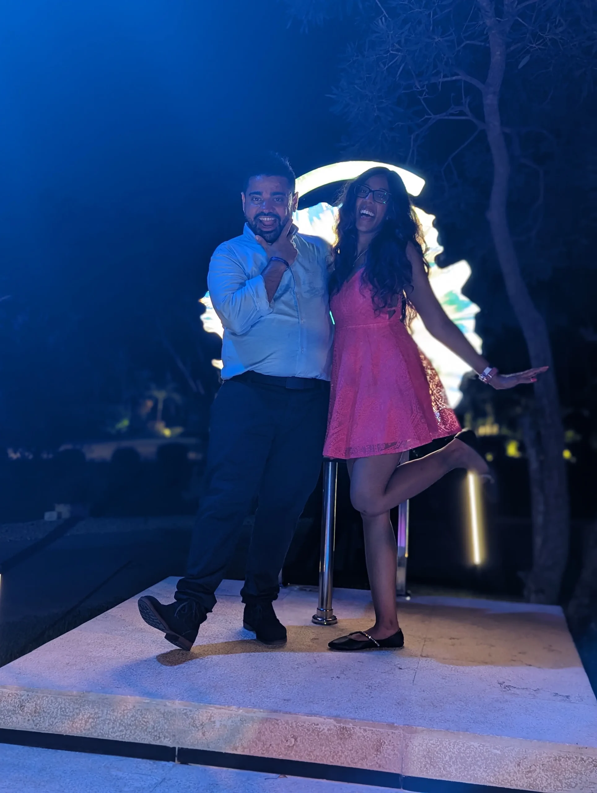 A playful nighttime photo of a couple posing on a lit platform. The man wears a light blue button-down shirt and dark trousers, resting his hand on his chin with a grin. The woman wears a bright pink lace dress and black flats, striking a pose with one leg lifted behind her. A bright, circular glowing light fixture creates a halo effect behind them against a dark, leafy background.