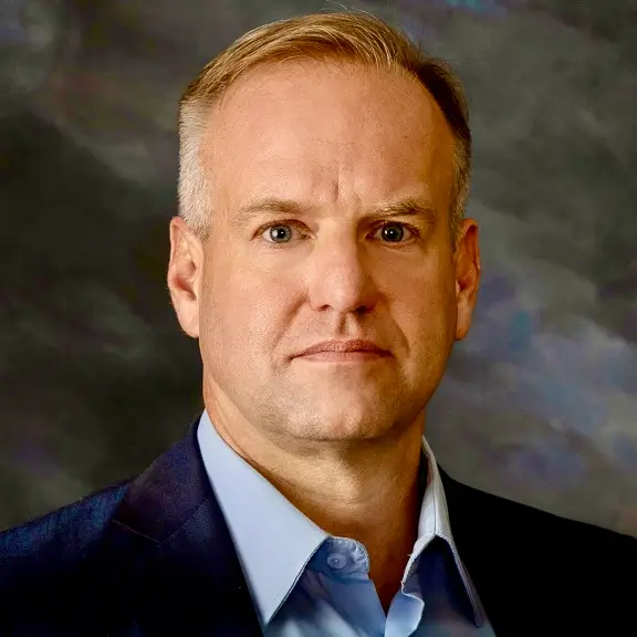 A professional headshot of a middle-aged man with short, thinning blonde hair and blue eyes. He is wearing a light blue button-down shirt under a dark navy blazer. He has a neutral, serious expression and is positioned against a mottled gray and blue studio background.