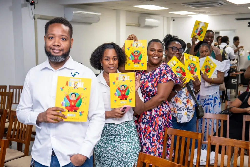 A line of smiling supporters, friends, and family holding up copies of the yellow children's book Dominic's Ausome World at a book signing event with author Giselle Trail McIntosh.