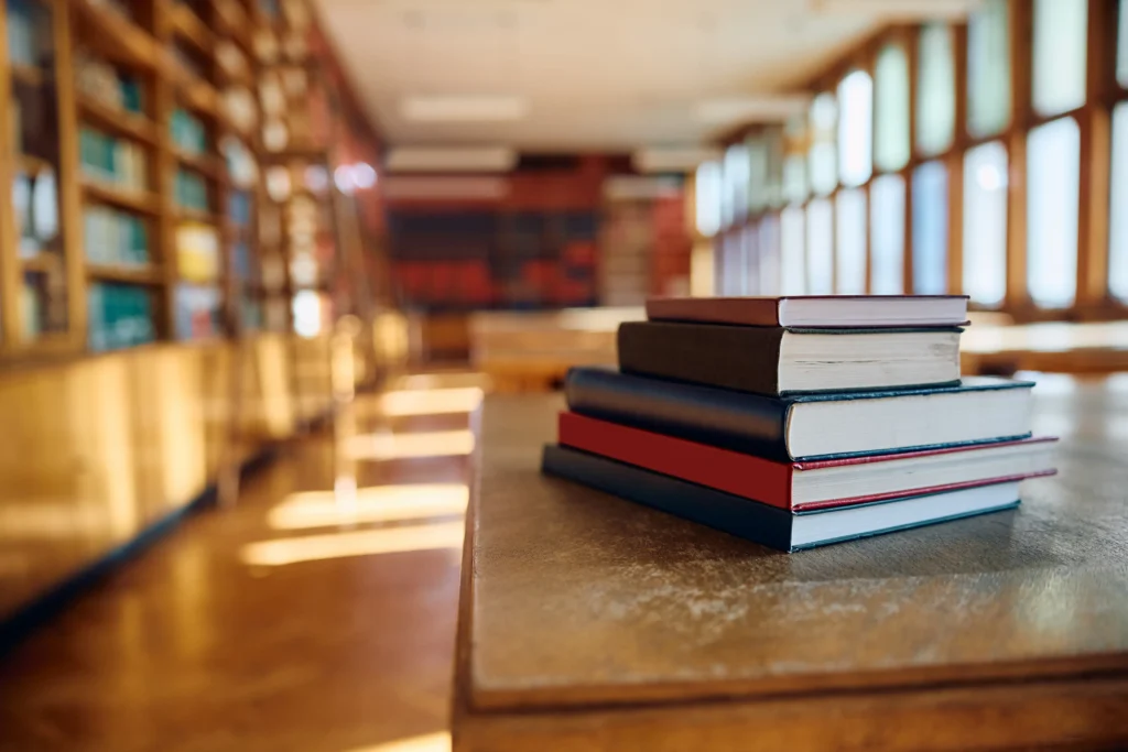 A close-up, slightly low-angle shot of a stack of five hardcover books resting on a polished wooden table. The background shows a blurred, sunlit library with tall wooden bookshelves and large windows casting soft light across the floor.
